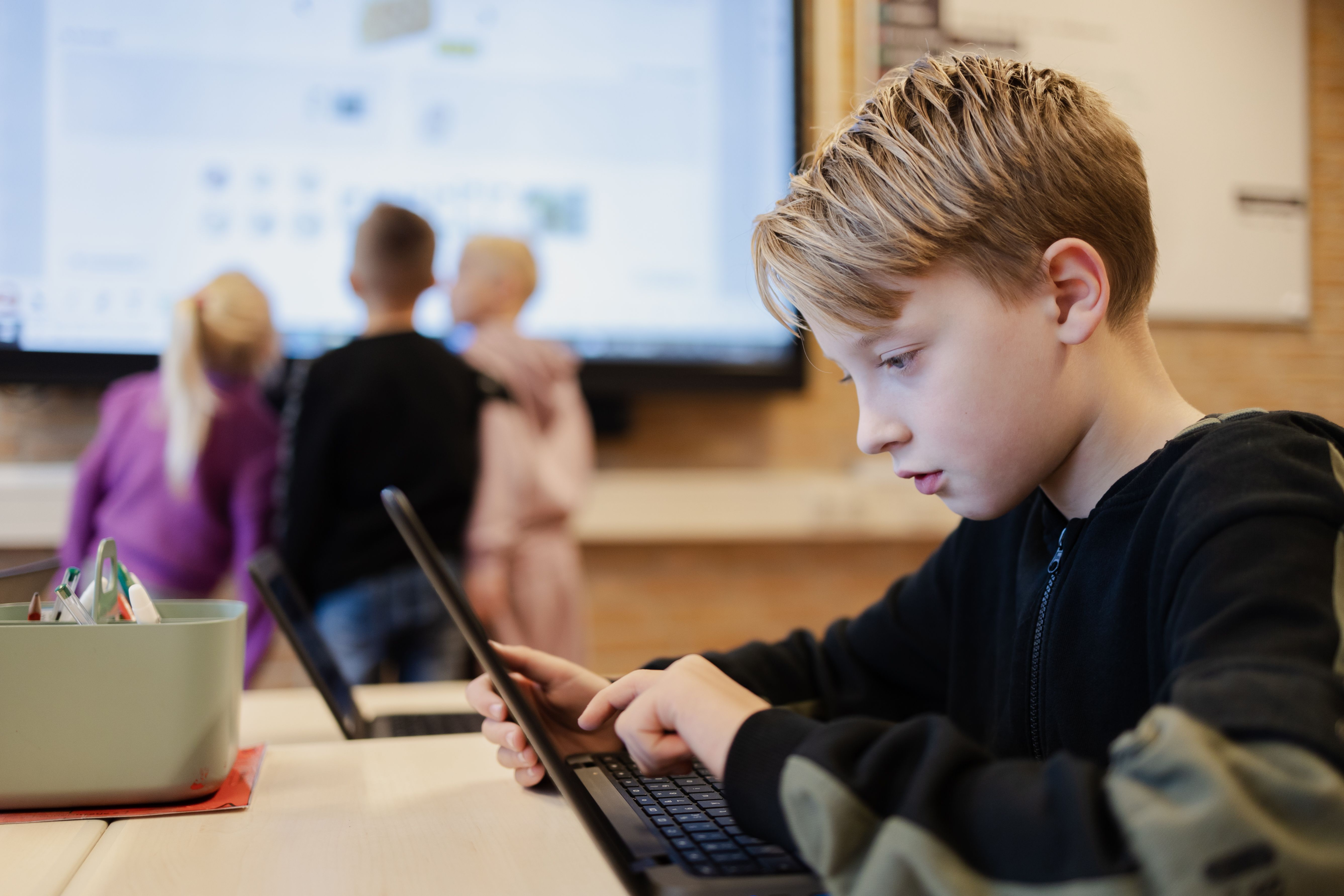 Leerling op chromebook met leerlingen bij het digibord in de achtergrond